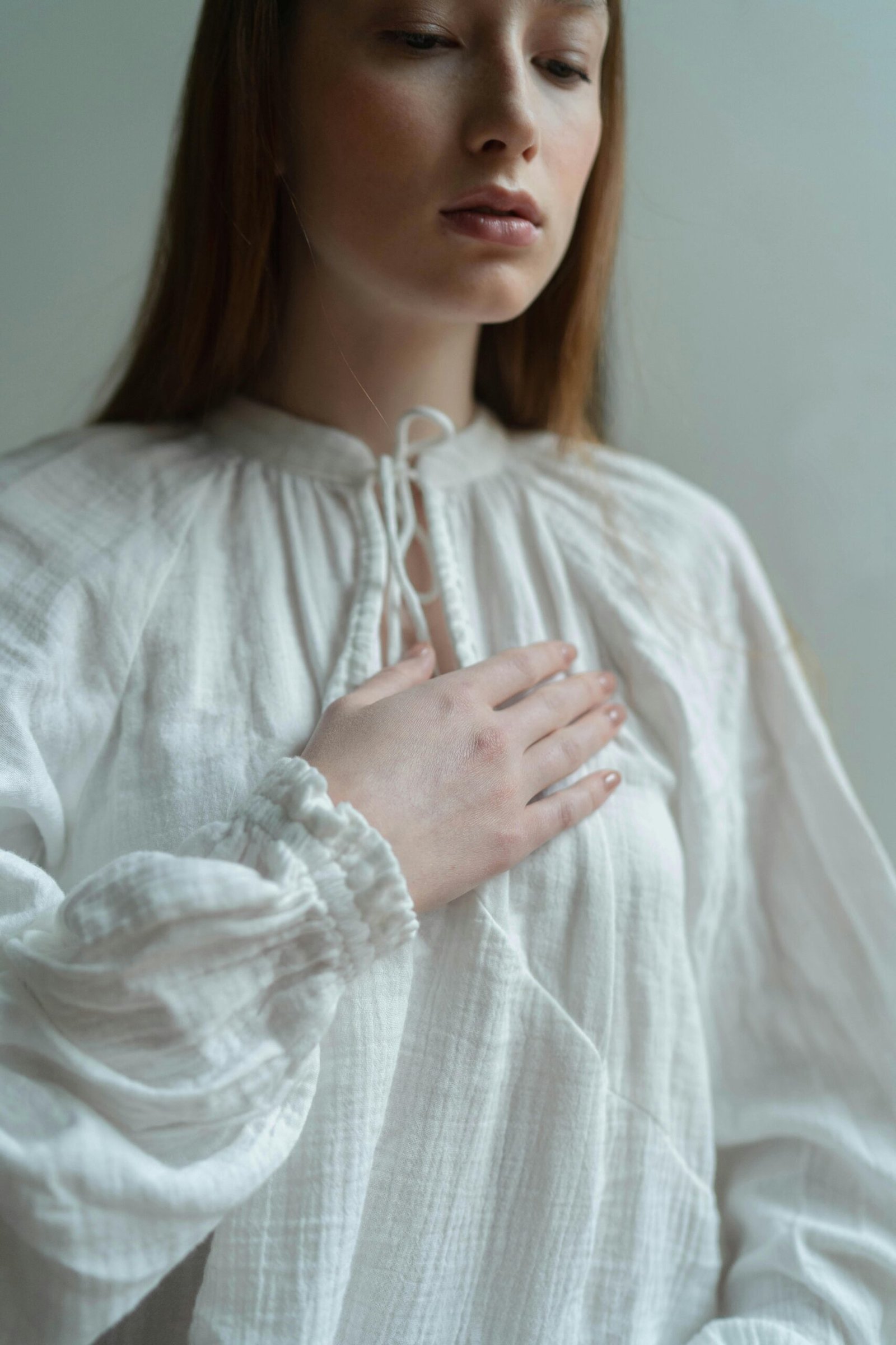 A contemplative woman in a white blouse gestures by placing her hand on her chest.