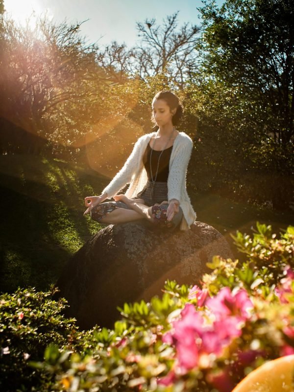 Woman in tranquil meditation pose outdoors, bathed in sunlight, surrounded by lush greenery.